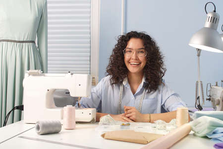 Dark haired tailor girl with glasses smiles and looks at camera in of home Studio interior