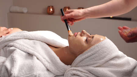 Beautiful woman with facial mask at beauty salon.Applying facial mask at woman face at beauty salon.Spa therapy for young woman receiving facial mask at beauty salon.Beautician does face mask.の写真素材