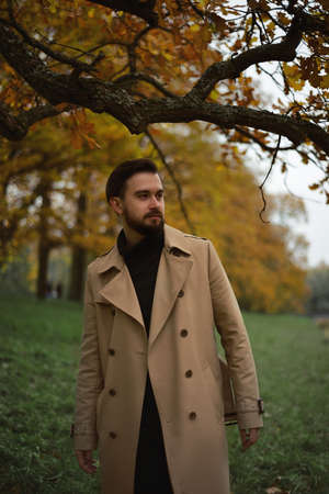A bearded dark-haired handsome man in a beige raincoat stands by a tree with yellow leaves, in the background an autumn forest.の写真素材