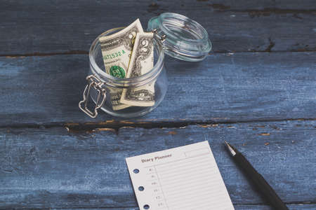 Two dollars in the jar and diary planner on a blue wooden background. Savings money or planning conceptの写真素材