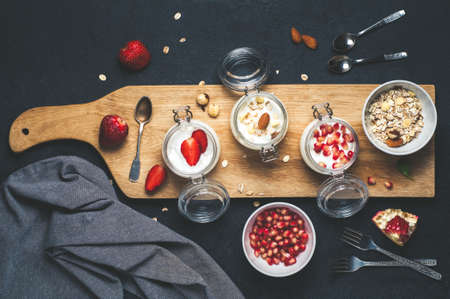 Homemade yogurt in glass jars, dry mix, pomegranate and strawberries on a wooden Board over dark concrete backgroundの写真素材