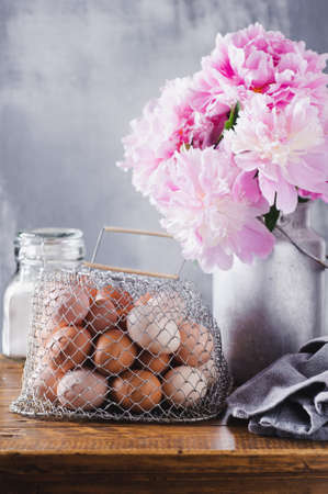 Concept of rural kitchen. Peonies in a can, flour and eggs on a wooden table against the background of a gray concrete wallの写真素材