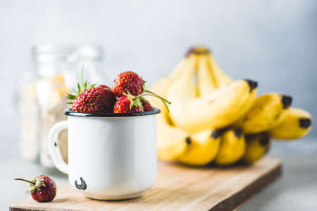 Fresh strawberries in a white metal cup, bananas on cutting board and cereals  over gray concrete backgroundの写真素材