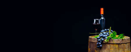 A glass of red wine, a bottle and a bunch of grapes with leaves on an old wooden barrelの写真素材