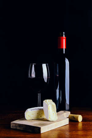 A glass of red wine, a bottle and a piece of cheese on a wooden table and a dark backgroundの写真素材