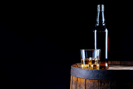 A glass of whiskey and ice cubes and a bottle on an old wooden barrelの写真素材