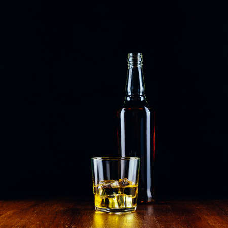 A glass of whiskey with ice cubes and a bottle on a wooden table and a dark backgroundの写真素材