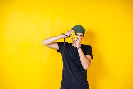 a young white man with a camera in his hands on a yellow background, a hipster in a black t-shirt...