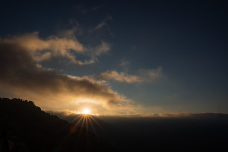 Fascinating panoramic view of the dawn in the mountains. A single large and obese cloud floats across the sky, through which the rays of the sun pass. Shadows from mountains and trees. High quality photoの写真素材