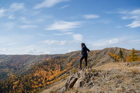 A girl in dark clothes walks along the top of the mountain, looks at the mountains and forest, the clear sky overhead, the autumn landscape. High quality photoの写真素材