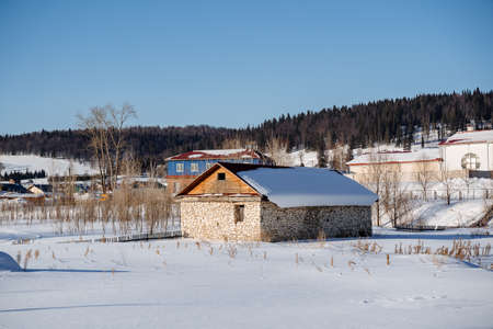 An stone house with a wooden fence in the middle of a snow-covered field on a sunny day. The roof is wooden and covered with snow. High quality photo. There are several more large houses nearby. High quality photoの写真素材