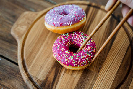Kitchen utensils made of wood. Unusual photo. Bright pink doughnuts lying on a round wooden plate are held with Chinese sticks. Plates made of natural material. High quality photoの写真素材