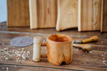 A homemade mug with an unusual handle stands on a wooden table, kitchen boards are exposed at the back, everything is made of wood. Nearby are tools for carving wood, sawdust everywhere. High quality photoの写真素材
