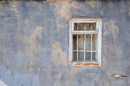 Old window with a wooden frame and a lattice in a purple wall with beige spots. Peeled white paint...
