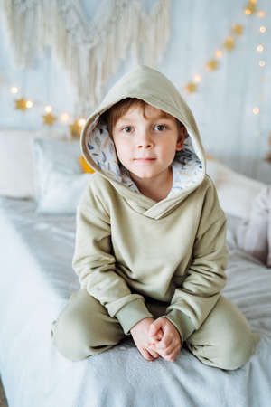 A boy in a light tracksuit sits on the bed. Behind him hangs a garland with stars. High quality photoの写真素材
