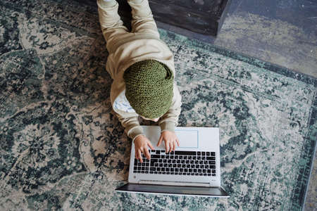 A boy in a knitted hat and a light suit lies on the carpet and practices in a laptop. The shot was taken from above. High quality photoの写真素材