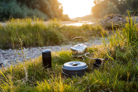 A close shot of a kettle and a flask by the river bank in the grass. Dinner in nature, meal of a tourist. Hike to nature to the river. Kettle, flask. High quality photoの写真素材