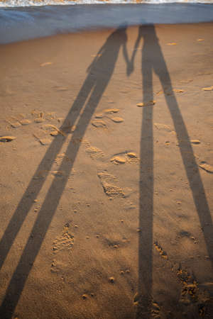 Long shadows on the sandy shore. A couple in love walks along the beach, holding hands. Relax by the sea, sea breeze and warm sandの写真素材