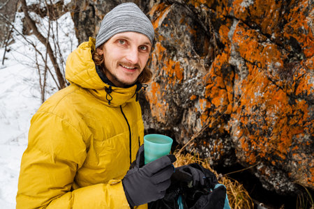An attractive and joyful man drinks tea on a rock. Picnic in the mountains. Travel in winter to the mountains. Adventures and exciting emotions. High quality photoの写真素材