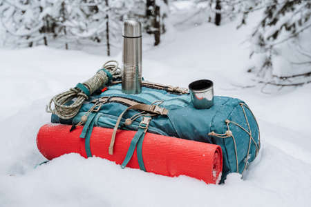 A tourist backpack lies on the snow, a red mat is fastened on the side, a bottle is next to it.