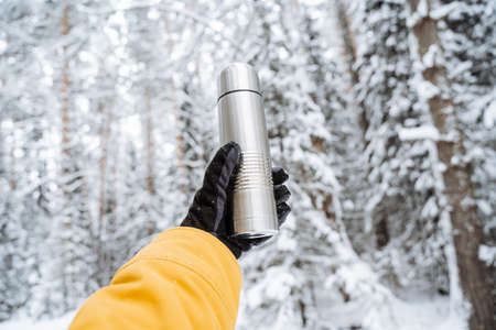Hand with a flask against the background of the forest in winter. Steel flask . Metal thermo. Snow-covered white forest, taiga in the snow. Outstretched arm. high quality photoの写真素材