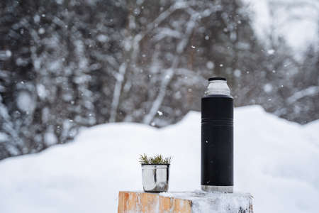 A black matte flask and a mug with a spruce twig stand on a snow-covered stump.Warm tea in the...