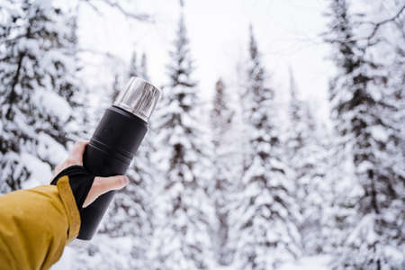 Black matte flask in hand. Metal lid and mug.Drink warm tea and coffee from on a walk in winter....