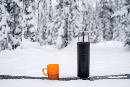 black matte flask and orange mug with coffee and tea in a snow. Hiking and picnic staff. Drinking warm tea in the winter frost. Minimalistic subject shot. high quality photoの写真素材
