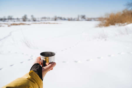 Thermal mug in hand, metal cup for hot tea. Camping on a clear sunny day, Breakfast against the backdrop of snow. high quality photoの写真素材