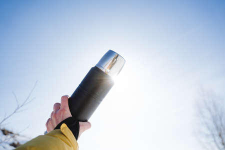 The hand holds a flask against the background of the sun, the concept of a thermo bottle with a hot drink, the glare of sunlight.の写真素材