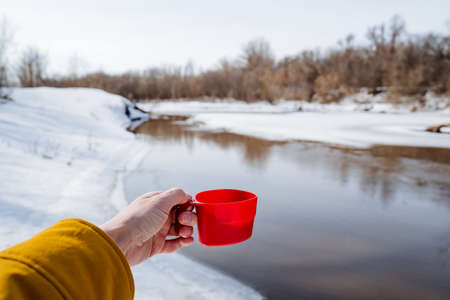 A red mug in a person's hand, a hand holding a glass against the background of the river, a tourist drinking tea in nature, a traveler winter hike, spring melting rivers. high quality photoの写真素材