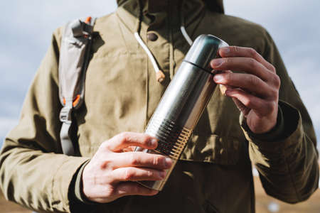 Vacuum silver flask is held in the hands of a man, the concept of a camping kitchen, a metal bottle for hot drinks, equipment for a hike. high quality photoの写真素材