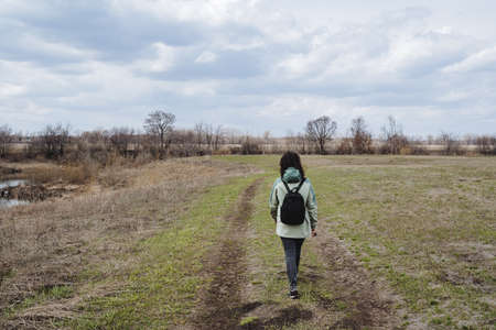 A girl with a backpack walks along the road in a green field, a spring walk through the park in nature, a hike in the forest in the spring. high quality photoの写真素材