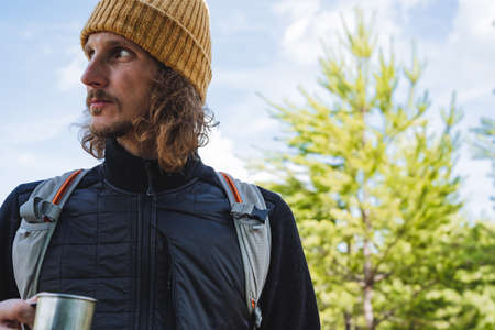 The guy looks away, the portrait of the man is close-up, the tourist holds a glass of tea in his hand, a journey through the mountains in the forest. A Caucasian young man is a hipster in a hat.の写真素材
