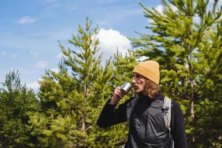 A guy drinking hot tea in nature holds a metal mug in his hand. A tourist in a yellow hat warms up with a hot drink. Morning at camp in the woods, have a glass of coffee.の写真素材