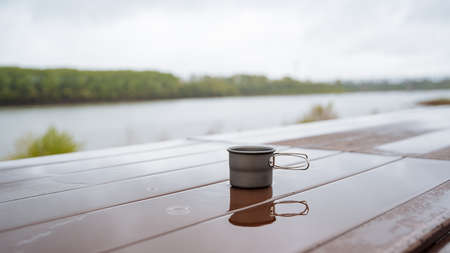 Coffee mug in nature, camping utensils, drinking hot tea on a hike, reflecting the silhouette of a glass in a puddle of water, a cup standing on the table wet, rain in the forest. high quality photoの写真素材