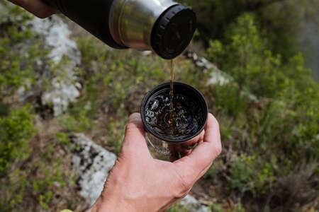 A man pours tea into a mug shot close-up, the concept of hot drinks in nature in the camp, a cup...