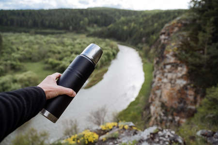 A flask in the hand of a person against the background of a river, hiking in the mountains, a...