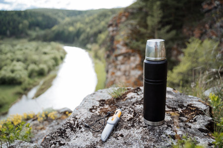 A flask stands on a stone against the background of the river, a landscape in nature in the mountains, a knife lies on a stone, the concept of a summer holiday in the mountains, hiking, hikingの写真素材