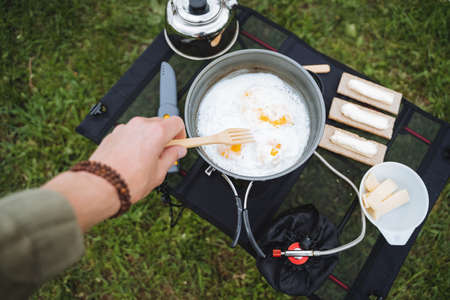 A guy roasts eggs in a frying pan at a campsite, cooking on a hike morning breakfast in nature, stir with a fork glaze, tourist utensils view from above. high quality photoの写真素材