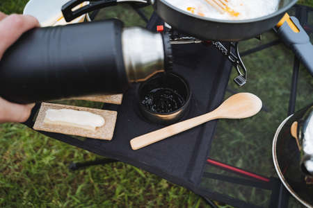 A hand holds a thermos pouring tea into a mug, a view from the top of the tourist utensils, outdoor...