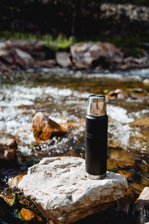 Thermo bottle to keep the heat inside, vacuum flask with coffee standing on a rock in the stream, mountain stream, metal vessel for liquid, tourist vacuum flask on a hike. high quality photoの写真素材