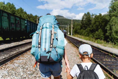 A mother and child go on a hike on the railway, a large blue backpack with things, a boy holds his mother's hand, an adult leads the child along the paths. high quality photoの写真素材