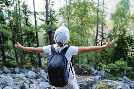 A boy on a hike raised his hands up, a child with a backpack looks to the sky, a hat protection...