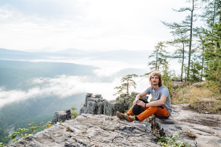 A tourist sits on a rock, a mountain trip alone on steep cliffs, morning fog in the mountains, a guy relaxing in nature. high quality photoの写真素材
