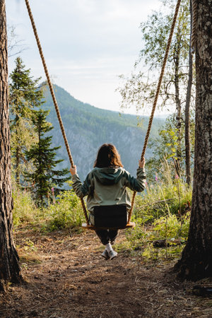 Swinging on a swing, The girl swings fascinatingly on a seat against the backdrop of mountains, a rope tied to a tree, a swing in the forest. high quality photoの写真素材