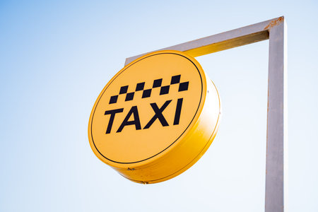 A taxi sign hangs against the sky, a taxi stop, a yellow banner with black text, letters in a circle, taxi checkers, sunlight. high quality photoの写真素材