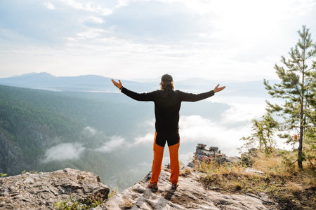 The sight of a shadi guy standing on a mountain looking forward hands to the sides, high in the mountains a happy man, a black jacket, morning dawn, hiking alone, the edge of the mountain.の写真素材