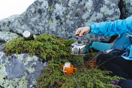 A tourist on a hike brews coffee in a glass, holds a metal kettle with boiling water in his hand, an orange mug stands on the grass, a juniper bush. high quality photoの写真素材