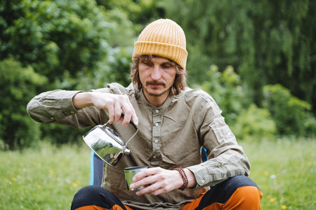 A hipster sits in nature drinking delicious tea, a guy in a hat pours a hot drink into a mug, a tourist has breakfast on a hike against the background of the forest, camping in a fieldの写真素材